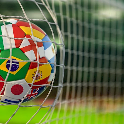 A soccer ball featuring various international flags is caught in a goal net on a blurred grassy field.