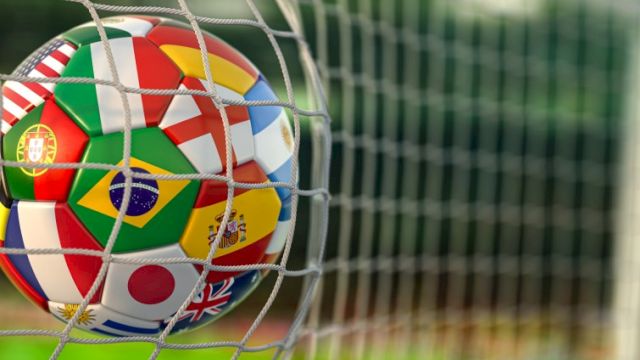 A soccer ball featuring various international flags is caught in a goal net on a blurred grassy field.