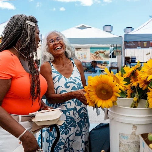 Two women smiling at a farmers' market with sunflowers; one holds a food container, while the other admires the flowers in a bucket.