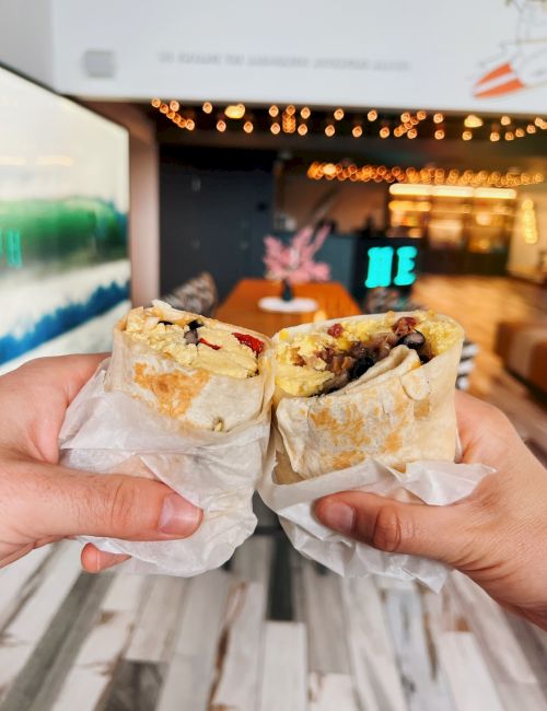 Two hands hold stuffed burritos inside a cozy, decorated room with string lights and a colorful painting on the wall.