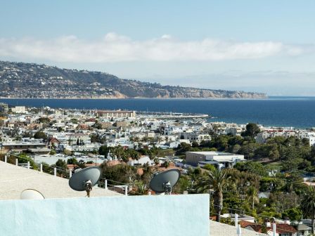 A coastal cityscape with buildings, greenery, and satellite dishes on rooftops; a vast ocean and distant hills in the background.