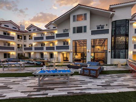 A modern hotel exterior at sunset with balconies, outdoor seating, and a ping pong table on a tiled patio surrounded by grass.