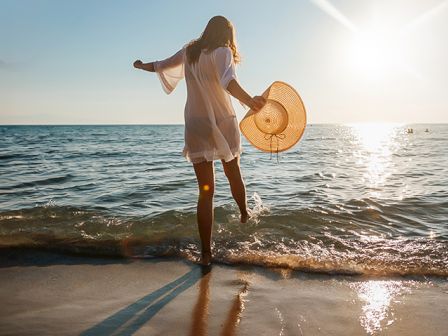 A person in a white dress and holding a hat is walking along a beach at sunset, with waves touching their feet.