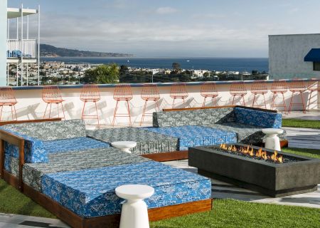 A rooftop lounge with blue seating, a fire pit, and ocean views. Pink bar stools line the railing; it's a relaxing outdoor space.