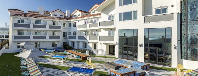 The image shows a modern hotel courtyard with outdoor seating, colorful tables, and a terrace, under a clear blue sky.