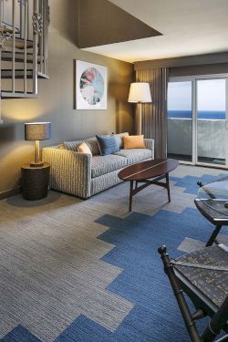 A cozy hotel suite with a living area, spiral staircase, blue zigzag carpet, sofa, coffee table, lamps, desk, and a balcony view.